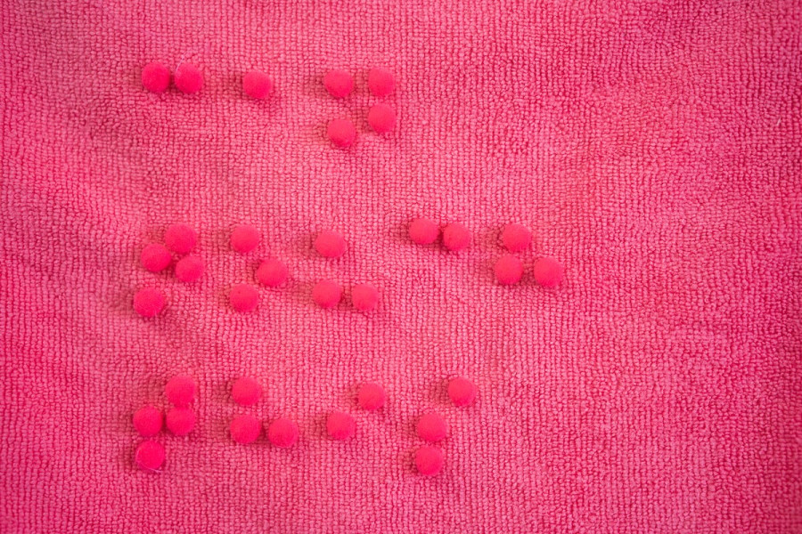Small, soft pink pom-poms arranged on a textured pink fabric surface to spell out the phrase 