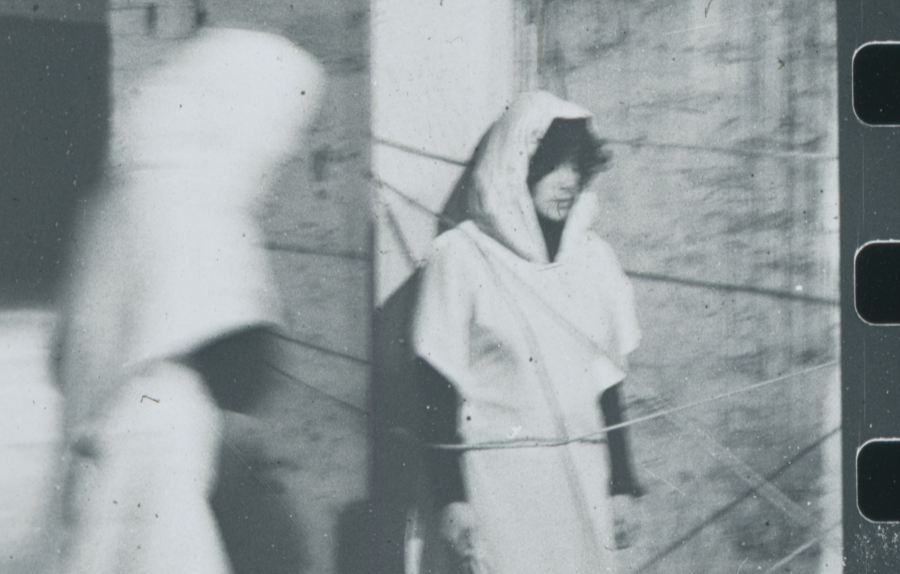 Two hooded figures in motion and stillness, connected by string in a dimly lit room, evoke mystery and quiet tension.